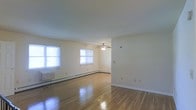 This single-floor space showcases a clean, understated design with a neutral color palette throughout. The main living area is highlighted by polished hardwood floors that reflect natural light, lending the room a warm and airy feel. Multiple windows along the exterior wall allow generous amounts of daylight to fill the space, enhancing its open atmosphere.

A ceiling fan with a light fixture is centrally positioned, providing both ventilation and ambient lighting. The walls are finished in a soft, off-white tone that complements the light wood flooring and contributes to the room's bright, welcoming character.

Baseboard heating units run along the lower portion of the walls, indicating a practical and efficient heating setup suited for year-round comfort. The layout is straightforward and open, offering flexibility for various furniture arrangements.

White trim and baseboards frame the space neatly, adding a subtle architectural detail that ties the room together. Overall, the space presents a functional, well-lit environment with classic hardwood flooring as its standout feature.