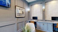 This thoughtfully designed business center lounge offers a sophisticated and functional environment for residents seeking a productive workspace. The room is defined by its refined architectural detailing, including full-wall wainscoting and paneling painted in a muted blue-gray palette that lends a calm, professional atmosphere throughout.

At the heart of the space is a built-in workstation wall featuring two individual computer setups, each framed by fluted pilasters and illuminated by warm brass wall sconces. The stations are set against a backdrop of glossy, handcrafted zellige-style tile, adding elegant texture and visual interest. Below the desk surface, soft blue cabinetry with brass hardware provides ample storage.

A comfortable lounge area anchors the center of the room, furnished with plush barrel chairs and a round wood accent table, offering a relaxed alternative to the dedicated workstations. A wall-mounted television displays local community content, adding a connected, informative touch to the space.

Near the window, a high-top bistro table with modern leather stools provides an additional casual seating option, bathed in natural light and framing views of a lush outdoor pool area. Framed artwork throughout the space adds a curated, gallery-like quality. A decorative niche with zellige tile and a live plant serves as a striking architectural focal point near the entrance, completing the space's polished and welcoming character.
