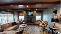 Located at 11217 18th Ave S in Tacoma, Washington, this single-floor community clubhouse is thoughtfully designed to blend rustic warmth with modern functionality. The lounge area serves as the heart of the space, showcasing exposed wood ceiling beams, shiplap-style wall paneling, and a dramatic stacked-stone fireplace with a mounted television above — framed by built-in seating and flanked by windows that bring in natural light. Comfortable sectional and accent seating arrangements make this an inviting gathering spot. Adjacent to the lounge, a spacious common area features a large communal dining table beneath stylish pendant lighting, reclaimed wood accent walls, and a dedicated coffee station. A fully equipped kitchen sits nearby, outfitted with dark walnut cabinetry, stainless steel appliances, open floating shelves, light quartz countertops, and a skylight that bathes the room in natural light. The space also includes a billiards room, visible from the main common area, providing a recreational outlet. A fitness area rounds out the amenities, equipped with cardio machines and free weights, with direct exterior access. Throughout the space, consistent use of mixed-material design elements — wood, stone, and neutral tones — creates a cohesive and welcoming atmosphere.