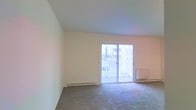 This single-floor residential space offers a clean, functional layout designed for comfortable everyday living. The main living area is a standout feature, characterized by light-colored walls and smooth ceilings that contribute to an airy, open atmosphere. Soft carpet flooring runs throughout, adding warmth underfoot and a quiet, comfortable feel to the space.

A prominent sliding glass door serves as both a design focal point and a practical feature, flooding the room with natural light and providing direct access to a private balcony. The balcony, framed by a white railing, overlooks the surrounding neighborhood, offering pleasant outdoor views and an inviting spot for fresh air.

Baseboard heating units line the walls, ensuring efficient climate control throughout the space. The overall color palette — soft whites and neutral tones — keeps the interior feeling bright and welcoming, while the uncluttered layout allows for flexible furniture arrangement to suit a variety of living styles.

The space presents a straightforward, well-maintained interior that prioritizes natural light, practical heating solutions, and seamless indoor-outdoor connectivity through its balcony access. It is well-suited for those seeking a simple, functional, and comfortable single-level living environment.