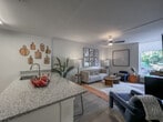 This single-floor residence in Williamsburg, Virginia offers a thoughtfully designed layout with a cohesive modern aesthetic throughout. The kitchen features sleek white cabinetry, granite countertops, light wood-style flooring, and a full suite of stainless steel appliances, including a range, microwave, refrigerator, and dishwasher — making it both functional and visually appealing. A striped runner rug adds a warm, casual touch to the galley-style layout.

The living area is styled with an eclectic mix of contemporary furnishings, including a plush sofa, bold navy accent chairs, and a curated gallery wall of framed line-art portraits. Large windows invite natural light and provide views of the surrounding greenery, enhancing the open, airy ambiance. A second sitting area features a wall-mounted TV, a navy dresser, and additional framed artwork.

The bedroom is a calm, restful retreat with light wood flooring, a ceiling fan, and large windows that bring in natural light. A classic wooden dresser with a gold octagonal mirror adds warmth and character to the space.

The bathroom is clean and well-appointed, with granite countertops, white cabinetry, light tile flooring, and tasteful decorative accents. Every room reflects careful attention to detail, creating an inviting and comfortable living environment.