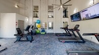Located at 1000 Country Oak Cir in Knoxville, Tennessee, this single-floor fitness center is housed in a striking space defined by soaring vaulted ceilings and dramatic verticality. Tall, decorative columns with a textured stone finish rise toward the ceiling, complemented by a large overhead ceiling fan that keeps the space well-ventilated. An upper-level balcony railing is visible above, adding architectural depth to the room.

Natural light pours in through multiple large windows along the walls, offering views of the surrounding greenery and community grounds. The patterned blue-toned carpet tiles add a modern, polished aesthetic throughout.

The cardio zone is well-equipped with multiple treadmills and stationary bikes, including upright and recumbent options, as well as elliptical cross-trainers. Three wall-mounted flat-screen televisions are positioned along one wall, ensuring an engaging workout experience. The strength and functional training area features a full dumbbell rack with a wide range of free weights, exercise benches, stability balls, resistance accessories, and floor space for stretching or bodyweight exercises. Wall-mounted mirrors are strategically placed to support proper form. This fitness center delivers a comprehensive and comfortable workout environment for all fitness levels.