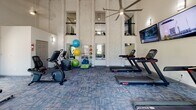 Located at 1000 Country Oak Cir in Knoxville, Tennessee, this single-floor fitness center is housed in a striking space defined by soaring vaulted ceilings and dramatic verticality. Tall, decorative columns with a textured stone finish rise toward the ceiling, complemented by a large overhead ceiling fan that keeps the space well-ventilated. An upper-level balcony railing is visible above, adding architectural depth to the room.

Natural light pours in through multiple large windows along the walls, offering views of the surrounding greenery and community grounds. The patterned blue-toned carpet tiles add a modern, polished aesthetic throughout.

The cardio zone is well-equipped with multiple treadmills and stationary bikes, including upright and recumbent options, as well as elliptical cross-trainers. Three wall-mounted flat-screen televisions are positioned along one wall, ensuring an engaging workout experience. The strength and functional training area features a full dumbbell rack with a wide range of free weights, exercise benches, stability balls, resistance accessories, and floor space for stretching or bodyweight exercises. Wall-mounted mirrors are strategically placed to support proper form. This fitness center delivers a comprehensive and comfortable workout environment for all fitness levels.