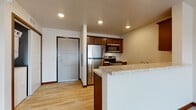 This single-floor residence offers a functional and comfortable layout designed for everyday living. The kitchen serves as a central highlight, featuring rich dark wood cabinetry, granite-style countertops, and a full suite of stainless steel appliances including a refrigerator, electric range, microwave, and dishwasher. Light wood-tone flooring runs throughout the kitchen and entry areas, adding warmth and easy maintenance. A breakfast bar with a stone countertop provides a natural transition between the kitchen and the open living area, which is finished with soft, neutral-toned carpet and recessed lighting overhead. The living space is generously proportioned, offering flexibility for various furniture arrangements. The bedroom is a comfortable retreat, featuring plush carpeting, a ceiling fan with integrated lighting, and a window that allows natural light and outdoor views. The bathroom is clean and straightforward, equipped with a full soaking tub and shower combination featuring a curved curtain rod, light wood-tone flooring, and crisp white finishes throughout. An in-unit laundry area adds a practical convenience to the space. Overall, the residence presents a clean, move-in-ready environment with consistent finishes and a cohesive neutral color palette throughout.