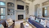Located at 3620 Huffines Blvd in Carrollton, TX, this thoughtfully designed single-floor space offers an impressive blend of classic architecture and modern amenities. The expansive main living area serves as the centerpiece, boasting soaring ceilings that lend a grand, open feel. A striking fireplace flanked by symmetrical arched built-in shelving and cabinetry anchors the room, while comfortable seating arrangements invite gathering and conversation. Natural stone tile flooring flows consistently throughout, lending warmth and cohesion to the entire space. Large, floor-to-ceiling windows in the dining area frame picturesque views of mature trees, manicured landscaping, and an outdoor fountain, bathing the interior in abundant natural light. The kitchenette is well-appointed with white shaker cabinetry, crisp white quartz countertops, subway tile backsplash, stainless steel appliances, open wood shelving, and a peninsula with bar seating — ideal for casual dining or refreshments. A quiet nook off the main area features built-in cabinetry and a cozy armchair, providing an intimate retreat. Recessed lighting, refined crown molding, and a carefully curated neutral color palette complete the polished, welcoming ambiance of this versatile space.