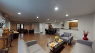 This well-appointed single-floor space offers a versatile, open layout designed to accommodate a variety of functions. Rich hardwood flooring flows consistently throughout, unifying the distinct functional zones and lending warmth to the overall aesthetic.

The main lounge area is generously proportioned, featuring multiple modern gray sofas, accent chairs, and a sleek coffee table — ideal for relaxed gatherings or informal meetings. Natural light filters in through sizable windows fitted with blinds, complementing the recessed ceiling lighting that keeps the space bright and welcoming. Classic accent pieces, including a carved wooden side table and decorative vases, add a touch of elegance.

A secondary lounge zone offers additional seating with leather sofas and armchairs, along with access to a glazed French door entry point. This area is well-lit by both recessed lighting and exterior-facing windows, creating an inviting atmosphere.

The conference and dining area centers around a large oval dark-wood table surrounded by leather chairs, accompanied by a counter-height bar area with stools — suitable for both formal meetings and social gatherings. A bookshelf with decorative accents completes this zone.

Adjacent to the main gathering areas is a functional kitchenette equipped with rich wood cabinetry, a sink, microwave, and a full-size refrigerator, providing convenient refreshment support for events or daily use.