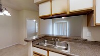 This single-story residence showcases an open floor plan that effectively merges the living, dining, and kitchen areas into a cohesive space. The kitchen is partially open, featuring a convenient pass-through window that maintains a connection with the living area while providing some separation. Granite countertops add a touch of elegance to the kitchen, complemented by a double-basin stainless steel sink and wooden cabinetry.

The living space is characterized by its openness, with a smooth transition from hardwood flooring to carpeting, delineating different functional areas. A three-light pendant fixture illuminates what appears to be the dining area, adding both style and functionality. The walls are painted in a neutral tone, creating a bright and airy atmosphere that's further enhanced by recessed lighting.

Vertical blinds on a large window suggest ample natural light during the day. The space is designed for modern living, with visible electrical outlets for convenience. The uncluttered layout offers flexibility in furniture arrangement and usage, making it adaptable to various lifestyle needs. Overall, the space presents a clean, contemporary aesthetic with a focus on openness and functionality.