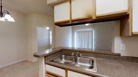 This single-floor space offers a practical and open layout that seamlessly connects the kitchen, dining, and living areas. The kitchen is equipped with a peninsula-style counter featuring dark granite-look laminate countertops and a double stainless steel sink, providing ample workspace and a convenient pass-through that opens directly to the main living area. White cabinetry lines the kitchen walls, complemented by warm wood-toned overhead cabinet trim, adding a touch of contrast. A vertical-blind-covered window allows natural light to filter into the kitchen, brightening the space. The dining area is defined by a classic chandelier with a multi-bulb fixture that casts a welcoming glow over the space. Soft, neutral-toned carpeting covers the living and dining areas, contributing to a comfortable, cohesive atmosphere throughout. The walls are finished in light, neutral tones that reflect light and enhance the open feel of the interior. Hardwood-style flooring at the kitchen entry transitions smoothly to the carpeted living space. Ceiling-mounted HVAC vents are integrated throughout, ensuring consistent climate control. Overall, the space presents a clean, functional layout well-suited for everyday living.