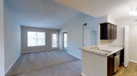 This single-floor residence showcases a thoughtfully designed open-concept layout that seamlessly connects the kitchen, dining, and living areas. The space immediately draws the eye upward with striking vaulted ceilings that lend a sense of airiness and volume throughout the main living area.

The kitchen is well-appointed with dark espresso cabinetry, stainless steel appliances including a refrigerator, microwave, and range, and a peninsula topped with light granite countertops that doubles as a casual dining and prep surface. A built-in sink on the peninsula adds functionality while maintaining the open sightlines into the living space.

The living area is generously sized and finished with plush wall-to-wall carpeting in a neutral tone that complements the soft grey and warm white wall palette. Large windows on the far wall allow abundant natural light to fill the room, while a glass-panel door provides direct access to a private balcony or patio, extending the living space outdoors.

A hallway leads from the main living area toward the bedrooms and additional rooms beyond, with continued carpeting and consistent finishes throughout. Ceiling-mounted track lighting and recessed vents are thoughtfully integrated, completing a clean, modern aesthetic ideal for comfortable everyday living.