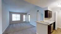 This single-floor residence showcases a thoughtfully designed open-concept layout that seamlessly connects the kitchen, dining, and living areas. The space immediately draws the eye upward with striking vaulted ceilings that lend a sense of airiness and volume throughout the main living area.

The kitchen is well-appointed with dark espresso cabinetry, stainless steel appliances including a refrigerator, microwave, and range, and a peninsula topped with light granite countertops that doubles as a casual dining and prep surface. A built-in sink on the peninsula adds functionality while maintaining the open sightlines into the living space.

The living area is generously sized and finished with plush wall-to-wall carpeting in a neutral tone that complements the soft grey and warm white wall palette. Large windows on the far wall allow abundant natural light to fill the room, while a glass-panel door provides direct access to a private balcony or patio, extending the living space outdoors.

A hallway leads from the main living area toward the bedrooms and additional rooms beyond, with continued carpeting and consistent finishes throughout. Ceiling-mounted track lighting and recessed vents are thoughtfully integrated, completing a clean, modern aesthetic ideal for comfortable everyday living.