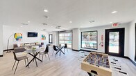 This contemporary community lounge is a thoughtfully designed single-floor space that blends social gathering and recreational amenities into one cohesive, open-plan layout. Light wood-look flooring runs throughout, complementing the crisp white walls and dark base trim that define the modern aesthetic.

The space is divided into distinct functional zones. A comfortable lounge area features a large sectional sofa in charcoal gray, set atop a bold, multicolored abstract area rug. A wall-mounted flat-screen TV and vibrant abstract artwork animate the walls, while a statement arc floor lamp and playful wall decals add personality and warmth.

Adjacent to the lounge, a casual dining and social zone offers multiple round tables with mixed modern seating, including woven accent chairs and sleek black side chairs, ideal for small group gatherings. A foosball table anchors the recreational corner, adding an energetic, playful element to the room.

A standout architectural feature is the full glass panel garage-style door that opens directly toward an outdoor pool area, flooding the interior with natural light and providing seamless indoor-outdoor connectivity. A large picture window further enhances the pool views. Recessed lighting throughout ensures the space remains well-lit at all hours. A teal accent wall and wood-paneled detail add pops of color and texture, completing this lively and inviting communal retreat.