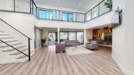 Located in Phoenix, AZ, this contemporary two-story commercial space offers a thoughtfully designed layout spread across two floors. The spacious main level welcomes visitors with soaring double-height ceilings, white shiplap detailing, and wide-plank wood-look flooring that runs throughout. A central reception counter with bar-height seating serves as a functional focal point, illuminated by a striking orb chandelier overhead. The open living area is anchored by a comfortable lounge arrangement and connects seamlessly to an outdoor patio through large sliding glass doors, flooding the interior with natural light. Private office spaces along the perimeter feature frosted glass partition walls and panel windows, providing both privacy and an open, airy feel. Exposed ductwork and a wall-mounted display screen add an industrial-modern character to the main floor. A sleek staircase with black cable railings leads to the upper level, which features patterned carpeting, vaulted ceilings with exposed beams, and a prominent skylight that bathes the space in natural light. A circular porthole window and sliding glass doors opening to an exterior deck further enhance the upper floor's brightness and connectivity to the outdoors. The overall ambiance is clean, modern, and welcoming.