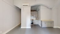 This unique 3-bedroom, 1-bathroom unit showcases a blend of character-rich architectural design and modern updates throughout its efficiently arranged single-floor layout. A defining feature of the space is the prominent barrel-vaulted arched ceiling in the main living and kitchen area, lending a distinctly historic and artistic quality to the interior. Smooth, polished concrete floors in a cool gray tone run continuously throughout, creating a cohesive, contemporary aesthetic.

The kitchen is thoughtfully appointed with gray shaker-style cabinetry, stainless steel appliances including a full-size refrigerator and range, a under-cabinet range hood, and a single-basin sink — all tucked neatly within the arched alcove. A built-in countertop with cabinetry extends into the adjoining space, offering additional storage and prep area.

The spacious primary bedroom benefits from its generous proportions, while the additional bedrooms are bright and airy, with one featuring a large window that brings in natural light and a wall-mounted mini-split system for individual climate control. Each room maintains the clean, minimalist palette of white walls and gray flooring.

The bathroom includes a vanity with gray cabinetry, a mirrored medicine cabinet, a tub-shower combination with white tile surround, and a warm parquet-style tile floor. Two closets provide practical storage, and a connecting hallway ties the layout together efficiently.