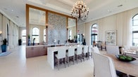 This expansive single-story space showcases a refined, contemporary design highlighted by soaring coffered ceilings and a series of dramatic floor-to-ceiling arched windows with sleek black frames. The abundant glazing floods the interior with natural light and frames lush outdoor views of palm trees, greenery, and what appears to be a waterfront setting.

The open-plan layout is anchored by a generous central kitchen island clad in white marble, surrounded by upholstered bar stools in crisp white. Behind it, a striking floor-to-ceiling herringbone mosaic tile feature wall is flanked by large mirrors and warm wood cabinetry, creating a bold focal point. An ornate multi-arm chandelier hangs above, adding a classic yet modern touch.

Flowing seamlessly from the kitchen area, the lounge spaces are thoughtfully arranged with comfortable neutral sofas, layered area rugs, and curated accent furniture. A built-in white entertainment wall with integrated cabinetry anchors one seating area, while high-top bistro tables offer additional casual seating throughout. Cream drapery panels soften the architecture, and large-format white tile flooring unifies the entire space, contributing to its bright, airy, and welcoming ambiance.