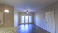 This single-floor residence offers a clean, modern aesthetic highlighted by rich, dark hardwood flooring that runs throughout the main living area, reflecting natural light and adding warmth to the space. The living room is an open, airy environment anchored by a set of elegant French doors that flood the interior with natural light while providing direct access to the outdoors. A ceiling fan with an integrated light fixture is centrally mounted, ensuring both comfort and functional illumination. The neutral-toned walls complement the overall design, creating a versatile backdrop suitable for a variety of interior styles. Granite countertops are visible at the edge of the living area, suggesting a connected kitchen space with quality finishes. White baseboards and trim provide a crisp contrast against the hardwood floors and soft wall tones. The layout feels open and unobstructed, offering a comfortable sense of space and flow. Recessed HVAC vents are integrated into the walls, maintaining the clean lines of the interior. Overall, the residence combines practical features with tasteful design elements, making it a well-appointed and inviting living environment.