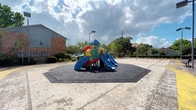 This outdoor recreational area is designed to serve as a multifunctional activity space. At its centerpiece sits a vibrant, multi-colored playground structure equipped with dual slides, climbing elements, and a covered canopy top. The structure is installed on a raised, octagonal rubber safety mat in dark grey, providing impact-absorbing protection around the play equipment.

The surrounding surface is a wide, open concrete area that extends to accommodate additional activities. A basketball hoop and backboard are visible toward the rear of the space, offering a dedicated area for sport. The grounds are bordered by mature trees and well-maintained shrubbery, contributing to a natural buffer and providing shade along the perimeter.

A brick building is positioned adjacent to the space, suggesting an institutional or community setting. Street lighting is present within the area, indicating the space can be utilized during evening hours. The open layout and variety of amenities make this a well-rounded outdoor space suited for active recreation across different age groups and activity types.