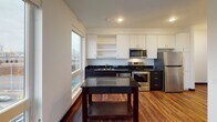 This well-designed single-floor unit offers a thoughtful open layout that maximizes both function and comfort. The kitchen is a standout feature, outfitted with a full suite of stainless steel appliances — including a refrigerator, electric range, and built-in microwave — complemented by dark granite countertops, a stainless steel sink, and a two-tone cabinetry scheme of white upper cabinets and dark lower cabinets. A freestanding kitchen island with a matching dark countertop provides additional prep space and storage. Rich hardwood flooring runs seamlessly throughout the entire unit, adding warmth and continuity. The living area is flooded with natural light from large, floor-to-near-ceiling windows that frame expansive views of the surrounding neighborhood and skyline, creating a bright and airy atmosphere. The bathroom is clean and functional, featuring a full soaking tub and shower combination, a dark-cabinetry vanity with a white basin, and grab bars for added accessibility. The 3D floorplan reveals a compact yet efficient layout with integrated murphy bed and shelving solutions in the living space, maximizing versatility. Overall, the unit presents a contemporary aesthetic with modern finishes and practical design throughout.