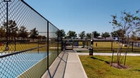 Located at 19101 Town Ridge Lane in Houston, TX, this outdoor amenity space offers residents a well-appointed recreational destination combining sport and pet-friendly facilities. The centerpiece of the area is a regulation pickleball court with a vibrant blue and green surface, fully enclosed by chain-link fencing and equipped with overhead lighting for extended play into the evening hours. A gated entry provides controlled access to the court, ensuring a safe and organized environment for players of all skill levels.

Adjacent to the pickleball court is a generously sized dog park, enclosed by sturdy black iron fencing with a secure double-gate entry system. The park features open green turf, a fire hydrant fixture, and benches, offering pets ample space to roam freely. Waste disposal stations are conveniently positioned near the entry points for ease of use.

The surrounding grounds are landscaped with neatly maintained grass and scattered mature trees that provide natural shade and visual appeal. Concrete pathways connect the various amenity zones and lead to nearby parking areas. A modern multi-story residential building frames the property, completing the community-oriented atmosphere. The entire space benefits from abundant natural light and open sky views, creating an inviting outdoor environment for both residents and their pets.