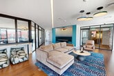 Located at 1 Green Island Boulevard in Worcester, MA, this thoughtfully designed amenity lounge spans a generous single floor, offering a sophisticated blend of modern aesthetics and functional gathering spaces. The main lounge area is anchored by a large sectional sofa set atop a bold, patterned area rug, complemented by designer pendant lighting and a striking teal accent wall featuring a built-in banquette nook. Warm hardwood flooring flows throughout, tying the open layout together seamlessly. A well-appointed wet bar area showcases floor-to-ceiling subway tile in both teal and deep blue, white quartz countertops, concrete-finish cabinetry, a built-in wine refrigerator, and pendant lighting — ideal for entertaining. A dedicated dining area, enclosed by sleek black-framed glass partitions, seats multiple guests around a live-edge wood table and is bathed in natural light from expansive black-framed windows overlooking the Worcester cityscape. A separate seating lounge with channeled leather armchairs offers additional space for conversation against a backdrop of panoramic city views. The space also connects to an outdoor rooftop terrace, furnished with lounge seating and bistro dining under string lights, providing sweeping views of the Worcester skyline.