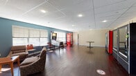 Located at 59 Broad St in Plattsburgh, New York, this single-floor communal space is designed to serve as a relaxed gathering and recreation area. The interior is finished with rich, dark hardwood-style flooring throughout, complemented by a drop ceiling with recessed lighting that provides bright, even illumination across the space.

The lounge area is defined by a bold teal accent wall that adds a pop of color against the warm cream-toned walls. Multiple windows fitted with roll-up shades allow natural light to filter in while offering views of the surrounding exterior. Comfortable upholstered armchairs and small side tables are arranged to encourage casual conversation and relaxation, alongside a wall-mounted flat-screen television.

A recreational corner features a classic pinball machine, adding an entertaining focal point to the space. Nearby, bistro-style round tables with bar-height chairs offer additional seating options. Along one section of the room, a row of vending machines — including beverage, snack, and specialty units — provides easy access to refreshments. A set of double glass doors serves as the main entry point, contributing to an open and welcoming feel. The overall ambiance is casual, functional, and well-suited for use as a shared amenity space.