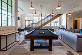 This dynamic community amenity space is designed for socializing, recreation, and productivity. The open-plan layout is anchored by polished concrete flooring and accented throughout by striking red pendant barn lights that lend a bold, industrial character to the interior.

The main lounge area features generous sectional sofas arranged around a cozy seating arrangement, complemented by a large-format wood-paneled accent wall mounted with multiple flat-screen televisions — ideal for group viewing. Adjacent to the lounge sits a full-size billiards table, perfect for casual entertainment. A dedicated card and game table with modern bucket chairs occupies a sun-drenched corner enclosed by floor-to-ceiling steel-framed windows, flooding the space with abundant natural light and offering views of the surrounding neighborhood.

A wet bar and kitchenette area lines one wall, featuring a white quartz countertop, stainless steel appliances, a sink, and dark cabinetry with mesh-front doors — all set against a richly textured reclaimed wood accent wall. A staircase with wood-and-steel detailing connects to additional levels of the building.

Tucked along one side of the space is a workstation nook equipped with desktop computers, offering a quiet corner for productivity. The overall ambiance blends modern comfort with an energetic, social atmosphere.