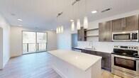 This single-floor unit showcases a thoughtfully designed open-concept layout with contemporary finishes throughout. The kitchen serves as the centerpiece, featuring rich dark-toned cabinetry paired with crisp white quartz countertops and a matching oversized island ideal for dining or meal preparation. A classic white subway tile backsplash complements the stainless steel appliances, which include a range, microwave, dishwasher, and refrigerator. A cluster of cylindrical pendant lights suspended from a track rail adds a modern, decorative touch above the island. Light hardwood flooring flows seamlessly throughout the unit, creating a warm and cohesive aesthetic. Large sliding glass doors in the living area invite natural light and provide direct access to a private balcony with outdoor views. The bedrooms are bright and airy, with windows framing natural scenery. Each bedroom connects to a well-appointed bathroom featuring dark wood vanities, white quartz countertops, undermount sinks, and tub-shower combinations. The overall ambiance is clean, modern, and move-in ready, with a neutral palette that offers versatility for any interior style.