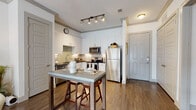This single-floor residence offers a thoughtfully designed layout that balances modern aesthetics with everyday functionality. The kitchen is a standout feature, outfitted with white shaker cabinetry, subway tile backsplash, stainless steel appliances including a refrigerator, range, and microwave, and a dark countertop that adds contrast. A freestanding high-top dining table with bar stools provides a casual space for meals and conversation. Warm hardwood-style flooring runs throughout the home, creating a cohesive and inviting atmosphere. The living area is bright and open, benefiting from tall ceilings, large windows, and a set of French doors that invite natural light and offer access to the outdoors. The bedroom is generously sized, featuring large windows dressed with curtains and blinds, providing both natural light and privacy. The bathroom is finished with a white vanity topped with dark granite, a subway tile accent wall, a large mirror with lighted fixtures above, and direct access to a well-organized walk-in closet with built-in shelving. Throughout the space, clean white walls, crown molding, and consistent flooring tie each room together into a cohesive, modern living environment.