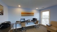 This single-floor space is designed as a functional and inviting business center, thoughtfully arranged to support focused work and productivity. The main workspace features a long, dark-toned desk that accommodates two full computer workstations, each equipped with a monitor and keyboard, making it well-suited for individual or shared use. Ergonomic rolling chairs in a warm tan finish provide comfortable seating at each station. The room is finished with soft blue-gray walls that create a calm, professional atmosphere, complemented by clean white baseboards and a neutral ceiling with recessed lighting that ensures consistent, even illumination throughout the space. A large, vibrant abstract canvas in warm amber and golden tones serves as a striking focal point on the main wall, adding a creative energy to the room. Natural light filters in through a sizable window fitted with white horizontal blinds, offering a connection to the outdoors while maintaining privacy. The flooring consists of a patterned commercial-grade carpet in earthy tones, adding texture and durability underfoot. Overall, the space strikes a balance between practical functionality and a welcoming, modern aesthetic.