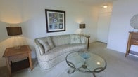 This single-floor residence offers a welcoming and thoughtfully designed living space with a clean, understated aesthetic. The living area serves as the centerpiece of the home, featuring plush wall-to-wall carpeting that adds warmth and comfort underfoot. A curved, floral-patterned sofa provides generous seating and lends the room a classic, elegant character, complemented by a round glass-topped coffee table with a decorative metal base. Soft ambient lighting is provided by a table lamp with a warm-toned shade, creating a cozy atmosphere. Framed wall art adds a tasteful decorative touch, enhancing the room's refined feel. The neutral color palette throughout — with light walls and soft floor tones — contributes to a bright and airy ambiance. An adjacent hallway visible from the living area suggests easy flow and connectivity to other areas of the home. The overall layout is practical and inviting, making efficient use of the available space while maintaining a comfortable and livable atmosphere suitable for everyday relaxation.