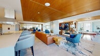 This single-floor communal space is thoughtfully designed to accommodate both relaxation and productivity. The main lounge area serves as the centerpiece, distinguished by a striking warm wood slat ceiling that adds texture and warmth throughout. A floor-to-ceiling brick fireplace anchors the space, functioning as a see-through feature that elegantly separates distinct seating zones. The lounge is furnished with plush blue sofas, cognac leather seating, and accent chairs arranged atop a fluid, marble-patterned area rug, creating a refined yet inviting atmosphere. Built-in wood shelving houses a large flat-screen television, while natural light pours in through expansive windows offering outdoor views.

Adjacent to the lounge, a glass-enclosed conference room provides a transparent yet private workspace, outfitted with a conference table, rolling chairs, a wall-mounted display, and a sculptural pendant light fixture. The kitchen is well-appointed with warm wood cabinetry, white quartz countertops, stainless steel appliances including a range hood, wall oven, and refrigerator, complemented by distinctive terrazzo flooring. Curated artwork and indoor plants add finishing touches throughout, contributing to the space's polished, contemporary character.
