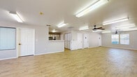 This single-floor space offers a versatile, open layout well-suited for a variety of uses, from community gatherings to office functions. The interior is finished with light wood-tone laminate flooring throughout, contributing to a clean, cohesive aesthetic across all areas.

The main open area is generously proportioned, featuring fluorescent ceiling lighting and a ceiling fan that together ensure consistent illumination and airflow. Multiple white panel doors provide access to separate rooms, including two ADA-accessible restrooms clearly marked with signage. A secondary open room at one end of the space benefits from natural light through multiple windows fitted with white horizontal blinds, offering views of surrounding greenery.

A dedicated kitchen area is fully equipped with stainless steel appliances, including a refrigerator, range, microwave, and dishwasher. White cabinetry with light wood trim lines the walls, complemented by light-toned countertops and a pass-through counter that opens toward the main gathering area, making it functional for serving or catering needs.

An additional room near the entrance features large windows and glazed sidelight panels on interior doors, allowing borrowed light to flow throughout. An illuminated exit sign marks the primary entrance, reinforcing the space's suitability for public or semi-public occupancy. Overall, the space presents a practical, well-maintained environment ready for immediate use.