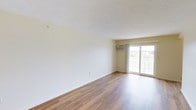 This single-floor space offers a clean and functional layout designed to maximize comfort and usability. The main living area is highlighted by warm, medium-toned hardwood-style flooring that runs throughout, providing a cohesive and inviting foundation for the space. Neutral, light-toned walls create a bright and airy atmosphere, offering a versatile backdrop for a variety of interior styles and furnishings. A large sliding glass door serves as a prominent architectural feature, flooding the room with an abundance of natural light while providing direct views of the surrounding greenery outdoors. Positioned above the sliding door is a wall-mounted air conditioning unit, ensuring year-round climate comfort. The textured white ceiling adds a subtle visual element while maintaining the overall simplicity and openness of the space. The room is generously proportioned, offering ample space for living and entertaining arrangements. The minimalist design and neutral color palette of this space make it highly adaptable, allowing occupants to customize the environment to their preferences with ease. Overall, the space presents a well-lit, functional, and welcoming interior with practical features throughout.
