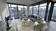 This single-floor residence is defined by its striking corner placement, where two full walls of floor-to-ceiling windows converge to deliver panoramic views of a dense urban skyline. The expansive glazing floods the living area with natural light throughout the day while framing an ever-changing cityscape as a dramatic backdrop.

The living room is thoughtfully appointed with a cohesive, contemporary design palette. A grey upholstered sofa anchors the seating arrangement, complemented by matching accent chairs that provide additional comfort without disrupting the room's clean aesthetic. A circular coffee table with a distinctive gold base and light stone top serves as a focal point, adding a warm metallic contrast to the otherwise neutral color scheme. A textured ivory area rug grounds the arrangement, adding depth and softness underfoot.

Track lighting mounted to the ceiling supplements the abundant natural light and provides targeted illumination for the space. Dark wood flooring adds warmth and contrast against the bright, airy atmosphere created by the glazed walls. The overall ambiance is modern and refined, balancing urban sophistication with comfortable, livable design.