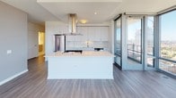 This single-floor high-rise unit showcases a contemporary design with an emphasis on natural light and panoramic city views. The open-concept living area is defined by expansive floor-to-ceiling windows that wrap around multiple sides of the space, framing sweeping views of the urban skyline and, from certain angles, Lake Michigan. Wide-plank gray hardwood flooring runs throughout the main living areas, complementing the clean, modern aesthetic.

The kitchen is a standout feature, centered around a large island with a gas cooktop and crisp white quartz countertops. Flat-front white cabinetry extends to the ceiling, providing ample storage, while stainless steel appliances—including a French door refrigerator and dishwasher—add a polished finish. A stainless steel island range hood serves as an architectural focal point above the cooktop.

The bedroom continues the floor-to-ceiling window theme, offering unobstructed city views and an abundance of natural light. The bathroom features large-format marble-look porcelain tile on both the floors and walls, a frameless glass walk-in shower with a built-in niche, and a floating wood-toned vanity with an undermount sink, delivering a refined, spa-like atmosphere throughout.
