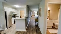 This single-floor residence offers a thoughtfully designed open-concept layout that connects the kitchen, living area, and bedroom through a central hallway. The kitchen is outfitted with white cabinetry, dark countertops, and stainless steel appliances, including a gas range with a ventilation hood. A peninsula with a built-in sink extends into the space, providing both additional workspace and a natural boundary between the kitchen and the rest of the unit. The hallway runs the length of the home, with warm wood-look flooring that flows consistently throughout, contributing to a cohesive and unified aesthetic. The walls are finished in a soft gray-green tone, complementing the clean white trim and baseboards. Framed artwork adorns the hallway wall, adding a curated touch to the space. The bedroom, visible from the hallway, is comfortably sized and warmly lit by a floor lamp. A seating area with a mustard-yellow accent chair is positioned along the hallway, offering a functional transition zone between the living spaces. Recessed and flush-mount ceiling lighting ensures the entire space is well-illuminated throughout.