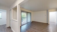 This single-floor unit is thoughtfully designed with a clean, modern aesthetic throughout. The living area features rich, dark hardwood-style flooring and is anchored by a full-width sliding glass door dressed with vertical blinds, opening to a private balcony with views of surrounding greenery. A pass-through opening connects the living space to the adjacent areas, enhancing the open feel of the layout.

The dining area continues the light hardwood-style flooring and is accented by a modest chandelier, lending a welcoming ambiance to the space. A doorway from the dining area leads directly to the bedroom, which is finished with soft, neutral carpeting and features a bright window with horizontal blinds that frame outdoor foliage, allowing ample natural light.

The galley-style kitchen is efficiently organized, featuring light gray cabinetry with contemporary bar-pull hardware, white countertops, and stainless steel appliances including a refrigerator and electric range. The bathroom is finished in crisp white tones, offering a full soaking tub with an integrated shower, chrome fixtures, and hardwood-style flooring. Overall, the unit maintains a consistent neutral palette throughout.