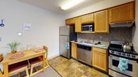 Located at 119 Dryden Rd in Ithaca, New York, this single-floor residential unit offers a practical and functional layout designed for comfortable everyday living. The open-concept living and kitchen area serves as the heart of the unit, where warm-toned carpet flows through the living space while stone-patterned tile defines the kitchen zone. The kitchen is well-appointed with stainless steel appliances, including a full-size refrigerator, dishwasher, gas range, and microwave, all complemented by honey oak cabinetry and a stone-tile backsplash. A compact dining area with a wooden table and seating sits conveniently adjacent to the kitchen. The living area provides ample space for relaxation, furnished with a sofa and armchair in deep burgundy tones, a wall-mounted flat-screen television, and a floor lamp. Large windows with horizontal blinds allow natural light to fill the room while offering views of the surrounding Ithaca streetscape. Multiple bedrooms are accessible from the main living space, each featuring closet storage and built-in shelving. The full bathroom includes a vanity with under-sink storage, a combination tub and shower with sliding glass doors, and a mirrored medicine cabinet. The unit also includes a wall-mounted HVAC unit for year-round climate control.