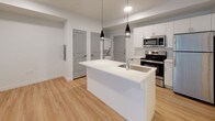 This well-appointed single-floor unit offers a clean, contemporary design throughout, characterized by crisp white walls, light wood-tone flooring, and a cohesive neutral color palette that lends the space a bright, airy feel.

The open-concept kitchen serves as the centerpiece of the layout, anchored by a large white waterfall-edge island with an integrated sink and pendant lighting overhead. White shaker-style cabinetry lines the walls, complemented by stainless steel appliances including a range, microwave, refrigerator, and dishwasher — providing a fully equipped cooking environment. The kitchen flows seamlessly into the living area, creating an inviting, connected space ideal for everyday living.

A notable convenience of this unit is the in-unit laundry, with a stacked washer and dryer tucked neatly into a dedicated alcove adjacent to the kitchen, keeping utility functions discreet and accessible.

The bathroom is finished in clean whites, featuring a full soaking tub with overhead shower, a white vanity with sink, and consistent light wood flooring that ties the space together with the rest of the unit.

The living area benefits from a large sliding glass door dressed with vertical blinds, providing access to an outdoor space while allowing natural light to filter through. The overall ambiance is modern, functional, and move-in ready.