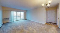 Located at 2121 W Main Rd in Portsmouth, Rhode Island, this single-floor residence offers a thoughtfully arranged layout with a clean, neutral aesthetic throughout. The living area is generously sized, finished with wall-to-wall carpeting and warm beige tones, and features large windows and sliding glass doors that open to a private balcony, filling the space with natural light and providing pleasant outdoor views. Baseboard heating runs along the walls, ensuring year-round comfort. A chandelier adds a touch of warmth to the room's ambiance. The kitchen is outfitted with white cabinetry, ample counter space, and a stainless steel sink, all set atop marble-style tile flooring that lends a clean, modern feel. The bedroom is spacious and carpeted, featuring two doorways — one leading to a well-organized walk-in closet with built-in shelving and another providing easy access to the hallway. The full bathroom is finished with the same marble-style tile flooring and includes a tub-shower combination with white tile surround, a toilet, a vanity with a white countertop, and a grab bar for added accessibility. The overall design is bright, functional, and move-in ready.