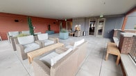 Located at 2710 N Proctor Street in Tacoma, Washington, this covered outdoor terrace offers a well-appointed communal lounge environment that blends indoor comfort with open-air surroundings. The space is finished with large-format light gray tile flooring throughout, lending a clean, modern aesthetic that complements the contemporary architectural detailing of paneled gray walls and warm terracotta-toned accent walls.

The terrace is generously proportioned, accommodating multiple distinct seating arrangements. Wicker lounge chairs and sofas with light gray cushions are grouped around natural wood coffee and side tables, creating comfortable social clusters suited for both casual gatherings and quiet relaxation. A built-in gas fireplace anchors one end of the space, framed by wicker armchairs and providing a cozy focal point. Large planters with ornamental grasses and topiaries add greenery and visual interest throughout.

Along the open perimeter, a wrought iron railing with brick pillar accents defines the edge of the terrace, offering elevated views of the North Proctor neighborhood and its tree-lined streets. Built-in bench seating along the railing provides additional spots to take in the outdoor scenery. Glass-paneled doors connect the terrace directly to the interior, ensuring seamless access between indoor and outdoor areas.