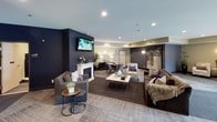 This single-floor common area serves as the social and entry hub of a modern residential building, thoughtfully designed to balance functionality with comfort. The entry foyer greets residents with large glass doors that flood the space with natural light, patterned carpet tiles, wall-mounted sconces, and convenient parcel lockers. An elevator provides seamless access to the rest of the building. Flowing from the foyer, the expansive lounge area is the centerpiece of the space. A bold navy blue accent wall serves as a dramatic backdrop for a wall-mounted flat-screen TV and a white-mantled fireplace below, creating a warm and inviting focal point. The seating arrangement includes plush gray sofas, patterned accent chairs, and sleek side tables, offering multiple conversation zones suited for both relaxed lounging and casual gatherings. A dedicated dining area with a round wood table and upholstered chairs provides a space for shared meals or informal meetings. Recessed lighting throughout maintains a bright yet cozy ambiance, while tasteful décor — including potted plants, decorative vases, and framed artwork — adds character. The overall palette of navy, taupe, and warm wood tones lends the space a polished, contemporary aesthetic.