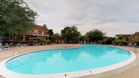 Carlton Apartments presents a welcoming community defined by its standout outdoor amenity space. The centerpiece of the property is a generously sized, kidney-shaped swimming pool with crystal-clear turquoise water, providing residents with an inviting space to relax and unwind. The pool area is finished with a wide concrete surround that comfortably accommodates lounge chairs and shaded seating areas beneath patio umbrellas, making it ideal for both leisure and socializing. Mature, leafy trees frame the perimeter of the outdoor space, offering natural shade and a sense of privacy, while well-maintained landscaping adds greenery and visual warmth to the setting. The surrounding brick residential buildings rise alongside the courtyard, contributing a classic, established architectural character to the community. The pool area benefits from an open-sky setting, allowing for ample natural light throughout the day. Overall, the outdoor common area at Carlton Apartments strikes a balance between functionality and comfort, providing a pleasant communal environment that serves as a central gathering point for residents. The combination of lush surroundings, classic brick architecture, and well-appointed poolside amenities gives the property a timeless, community-oriented ambiance.