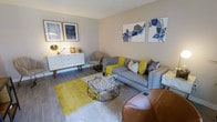 This single-floor living space showcases a thoughtfully designed interior with a cohesive mid-century modern aesthetic. The living area serves as the focal point, finished with light grey laminate flooring that runs throughout and a textured ceiling that adds subtle depth overhead. Neutral warm-beige walls provide a versatile backdrop, allowing the carefully chosen furnishings and accents to stand out.

The seating arrangement centers around a grey upholstered sofa adorned with a mix of yellow, white, and patterned throw pillows, complemented by a cognac leather accent chair that introduces a rich, warm contrast. A geometric black wire coffee table sits atop a bold mustard-yellow area rug, anchoring the space with a striking visual element. A patterned accent chair in the corner adds textural variety to the arrangement.

Along one wall, a sleek white mid-century sideboard with tapered wooden legs provides functional storage while reinforcing the design theme. Decorative items, including a brass table lamp and small framed artwork, are styled atop it. Two sets of framed wall art — one featuring abstract blue patterns, the other geometric blue and neutral shapes — complete the polished, gallery-like feel of the room.