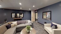 This well-appointed single-floor space combines comfort and modern design through a cohesive navy blue and gray color palette that flows throughout. The main lounge area serves as the centerpiece, featuring a white fireplace framed by deep navy accent walls, recessed lighting, and a wall-mounted television. Plush gray sectional seating and cream accent chairs are arranged around a round wood coffee table, creating a welcoming and functional gathering space. Framed artwork adds personality and visual interest throughout.

Adjacent to the lounge is an intimate dining nook, defined by a tray ceiling with a bold navy border detail. A round wood table surrounded by upholstered chairs offers a comfortable setting for smaller gatherings, complemented by greenery and curated wall art.

The kitchen is a standout feature, boasting warm honey-toned cabinetry, granite countertops, and stainless steel appliances including a gas range. A generous island with pendant lighting provides additional prep and serving space, while light hardwood-style flooring adds warmth and contrast to the bold wall tones. The open sightlines connecting the kitchen to the dining and lounge areas make the overall layout ideal for entertaining and everyday use.
