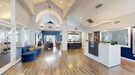 This single-story community or leasing office space offers a generously scaled, open layout thoughtfully designed to welcome visitors and foster a polished, professional atmosphere. The architecture immediately impresses with dramatically vaulted ceilings supported by exposed beams and adorned with a statement sputnik chandelier, creating a grand sense of vertical space throughout the main area.

Large arched windows and French doors line the walls, flooding the interior with abundant natural light and offering views of the surrounding outdoor greenery. The warm hardwood-style flooring flows continuously throughout, unifying the various functional zones. A reception area with a sleek white counter provides an organized point of entry, while a dedicated lounge area features curated furnishings in deep navy, gray, and amber tones, anchored by a textured accent wall with a wall-mounted television and wood credenza.

A bold navy-painted seating nook with a dining table and mixed-material chairs offers a more intimate setting framed by arched windows and French doors. A compact refreshment station includes a commercial coffee machine, stainless steel appliances, and white cabinetry with subway tile accents. Together, these elements create a sophisticated, inviting, and highly functional communal environment.