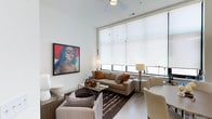 Located at Main & Clay, this single-floor residence blends contemporary design with thoughtful functionality. The living area is an immediate standout, boasting impressively tall ceilings, a ceiling fan, and a wall of large windows dressed with roller shades that flood the space with natural light while offering views of the surroundings. Bold artwork adorns the crisp white walls, adding a vibrant artistic energy to the room. The living space is anchored by a neutral sofa accented with colorful throw pillows, a glass coffee table, and a patterned area rug, creating an inviting and stylish atmosphere.

The kitchen is efficiently designed with white cabinetry, a grey subway tile backsplash, light wood-toned flooring, and a full suite of stainless steel appliances including a dishwasher, range, microwave, and refrigerator. A compact dining area with a round table and white chairs sits adjacent, complemented by a decorative mirror on a concrete accent wall.

The bedroom offers a serene retreat with warm, soft lighting, a well-appointed bed with neutral linens, a wall-mounted sconce, and tasteful artwork. The bathroom is clean and practical, featuring light wood-tone flooring, a white vanity, and a glass-enclosed walk-in shower, completing this well-designed urban residence.