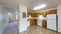 This two-story residence presents a practical and well-organized layout designed for comfortable everyday living. The kitchen serves as a central feature of the main level, outfitted with warm honey-toned wood cabinetry that lines the walls in an L-shaped configuration, providing generous storage space. White appliances — including a refrigerator, electric range with overhead ventilation, and dishwasher — complement the neutral countertops and contribute to a clean, cohesive look. The flooring features a durable stone-patterned tile, well-suited for a high-traffic cooking area. A utility panel is conveniently mounted on the adjacent wall. Overhead, a flush fluorescent light fixture illuminates the workspace, while a textured ceiling adds subtle visual character throughout. A doorway from the kitchen offers a glimpse into an adjoining carpeted living area, which benefits from natural light via a nearby window. The two-floor layout provides clear separation between the living and private areas of the home, enhancing both functionality and privacy for occupants.