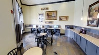 This single-floor business center offers a functional and well-organized environment suited for work and productivity. A row of computer workstations lines one wall, each equipped with monitors and ergonomic office chairs, providing ample space for individual use. A dedicated printing and copying station is positioned nearby, adding convenience for document management tasks.

The flooring throughout the space features slate-tiled stonework in earthy tones, lending a grounded, professional aesthetic that contrasts warmly with the light cream-colored walls. Dark crown molding frames the upper perimeter of the room, adding a refined architectural detail. A series of framed artwork in neutral and earth tones is arranged along the walls, contributing to a polished yet welcoming ambiance.

Casual seating, including round bistro-style tables and black wooden chairs, is arranged in the central area, offering a relaxed alternative workspace or meeting spot. Natural light filters in through a window on one side, brightening the room. Recessed ceiling lighting ensures consistent illumination throughout the space, making it equally comfortable during evening hours.