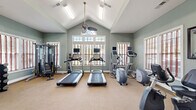 This single-floor fitness center offers a thoughtfully designed workout environment housed beneath a dramatic vaulted ceiling that creates an open, airy atmosphere. The architectural centerpiece is the steeply pitched ceiling, which peaks at the center of the main exercise area and is complemented by ceiling fans that promote air circulation throughout. Large windows with louvered shutters line the walls, including a prominent arched transom window at the front, filling the space with natural light and providing outdoor views.

The main workout area is carpeted and features a comprehensive selection of cardio equipment, including multiple treadmills and elliptical machines, each equipped with individual display screens. A multi-station strength training machine, free weights, and a dumbbell rack are positioned along one wall, while exercise balls, resistance equipment, and yoga mats are organized in a dedicated functional training zone.

Adjacent to the main floor is a smaller secondary room, accessible through glass-paneled French doors, which appears to serve as a children's area or small lounge space. A mini-refrigerator and wall-mounted shelving add practical utility to the facility. The sage-green walls, abundant lighting, and generous floor area collectively create an inviting and functional fitness environment.