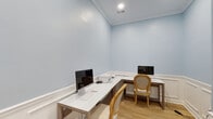 This business center in Horn Lake, Mississippi, is a compact, single-floor workspace designed with both functionality and refined aesthetics in mind. The room is finished in a soft, calming light blue paint above crisp white wainscoting with raised panel detailing that lines the lower walls, creating a polished and professional atmosphere throughout.

The space is equipped with an L-shaped white desk configuration supported by natural wood-toned frames, providing ample surface area for two workstations. Each station features a desktop computer and keyboard, while a shared printer rests at the corner junction of the desks. Elegant upholstered round-back chairs in a classic style complement the room's refined character.

Light hardwood flooring runs throughout, adding warmth and continuity to the space. Crown molding accents the ceiling, further elevating the room's traditional design sensibility. Recessed lighting provides even, functional illumination overhead.

A standout architectural feature is the glass-paned French door, which allows natural light to filter in from an adjoining area while maintaining a degree of privacy. The door's multi-lite design and white trim are consistent with the room's overall traditional style.

Overall, this business center offers a welcoming and well-equipped environment well-suited for productivity, combining classical design elements with practical workstation amenities.